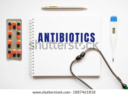 ANTIBIOTICS is written in a notebook on a white table next to pills and a stethoscope. Medical concept