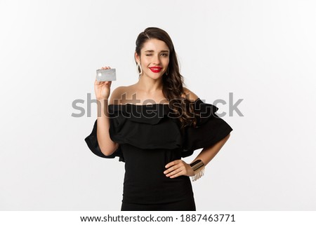 Beauty and shopping concept. Fashionable woman in black dress, wearing makeup, winking at camera and showing credit card, standing over white background