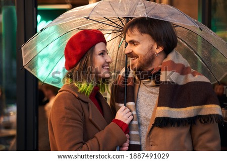 Waist up portrait view of happy woman strolling with her man. They are hiding under umbrella with joy. Smiling couple is looking at each other with tenderness
