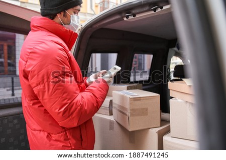 Side view of the courier wearing protective mask holding smartphone while scanning a barcode. Man taking out parcel boxes from the car while making delivery. Stock photo