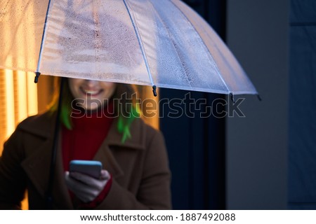 Outdoor picture of smiling positive model holding umbrella and smartphone, having peaceful facial expression, using her device actively, spending time alone. Youth and technology concept