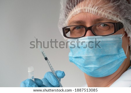 Coronavirus vaccine concept. Medic, doctor or nurse with covid-19 vaccine vial and syringe. Mature European female tech in face mask, glasses, disposable hat, blue gloves looks at camera.
