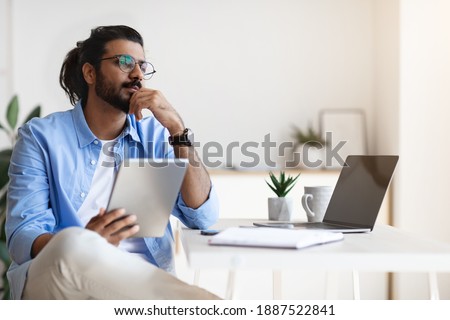 Pensive Young Arab Male Freelancer Using Gigital Tablet And Daydreaming At Home, Handsome Eastern Man Thinking About New Business Ideas, Sitting At Desk Touching Chin And Looking Aside, Free Space Royalty-Free Stock Photo #1887522841