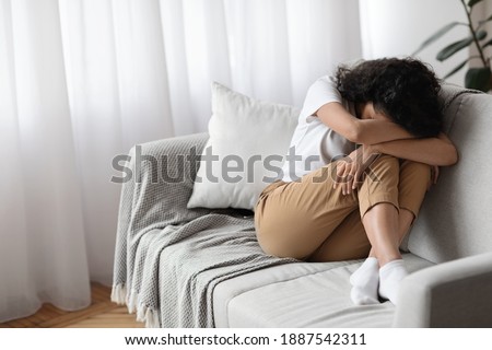 Depressed brunette woman sitting on couch at home alone, crying, suffering from loneliness during lockdown, copy space. Upset young lady having bad day, spending weekend on her own, feeling lonely Royalty-Free Stock Photo #1887542311