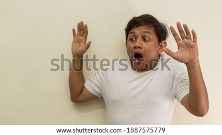 Wow and shocked face expression of young Asian Malay man in white t-shirt with both hand upward looking on empty space on isolated white background. Selective focus.