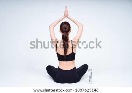 A young healthy girl, in a black top and leggings, practicing yoga, sits in a yoga pose, turning her back, a bottle of water stands next to her. Yoga lady on a white background. Copy space.