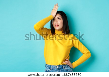 Emotions and lifestyle concept. Annoyed asian girl roll eyes and face palm, standing bothered in yellow pullover against blue background. Royalty-Free Stock Photo #1887658780