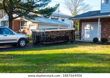 A small short beat up black and white dumpster containing wood debris located in a driveway in front of a garage door Royalty-Free Stock Photo #1887664906