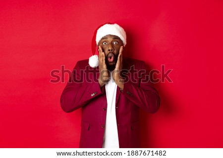 Christmas, party and holidays concept. Shocked Black man in santa hat reacting to new year offer, standing against red background