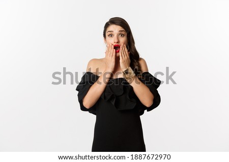 Fashion and beauty. Image of attractive female model in black dress reacting to announcement, looking amazed at camera, standing surprised over white background