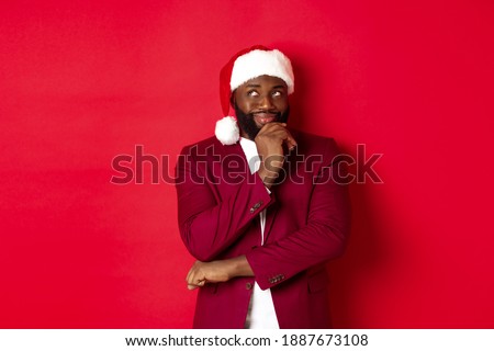 Christmas, party and holidays concept. Smiling african american man making new year plans, looking thoughtful at upper left corner, wearing santa hat, red background