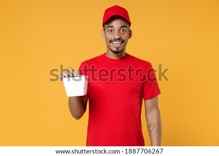 Delivery employee african man 20s in red cap blank print t-shirt uniform work courier service concept hold Chinese food cuisine and takeaway carton container box isolated on yellow background studio