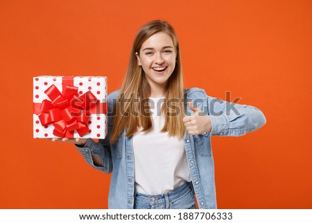 Cheerful young woman 20s in casual denim clothes isolated on orange background. Valentine's Day, Women's Day, birthday, holiday concept. Hold red present box with gift ribbon bow, showing thumb up