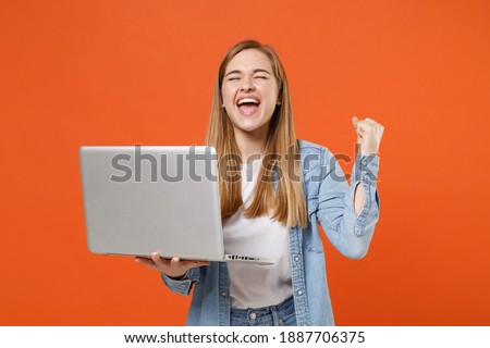 Happy young woman in casual denim clothes posing isolated on orange background studio portrait. People lifestyle concept. Mock up copy space. Hold laptop pc computer, doing winner gesture celebrating