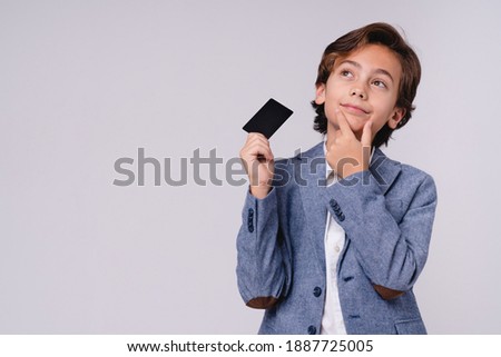 Thoughtful young boy with credit card in formal attire isolated over grey background