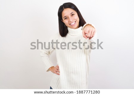 Young brunette woman wearing white knitted sweater against white background  pointing at camera with a satisfied, confident, friendly smile, choosing you