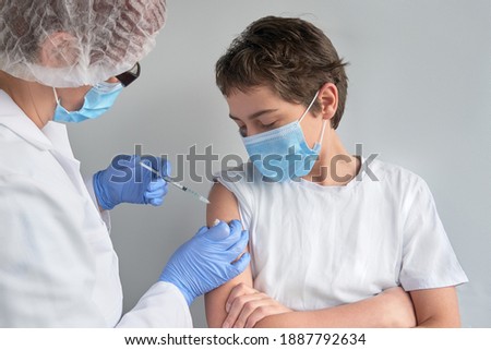 Coronavirus, flu or measles vaccine concept. Medic, doctor, nurse, health practitioner vaccinates teenage boy with vaccine in syringe. She is wearing uniform, hut and gloves. Both have face mask Royalty-Free Stock Photo #1887792634