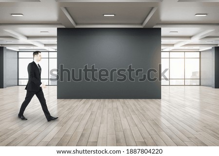 Businessman walking in gallery interior with empty black wall, city view and daylight. Concrete floor. Mock up