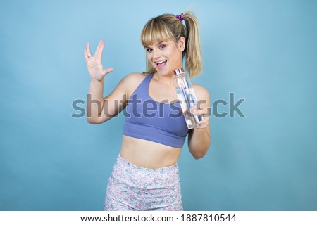 Young beautiful woman wearing sportswear holding a bottle of water over isolated blue background doing hand symbol