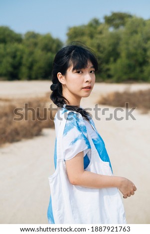 Black hair French braid girl in white and blue tie dye standing in front of mangrove forest with canvas bag. Royalty-Free Stock Photo #1887921763