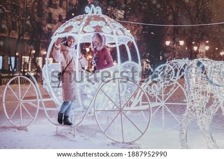 Girl friends taking selfie photo in illuminated fairy tale carriage with horses decorated with lights and garlands for Christmas night and New Year holiday. Friendship and winter vacation concept