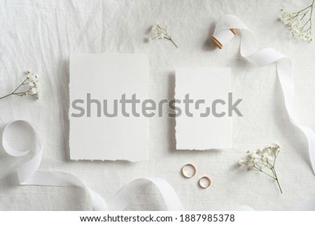 Blank wedding invitations cards mockups with space for text, golden rings, ribbon flowers on white textile background. Flat lay, top view.