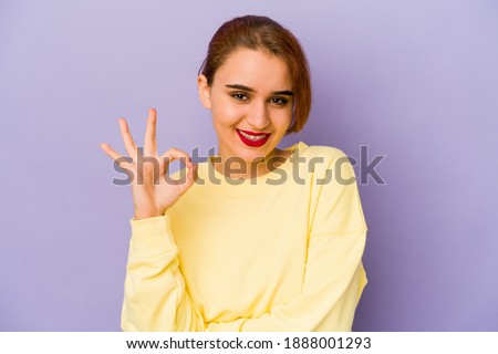 Young arab mixed race woman winks an eye and holds an okay gesture with hand.
