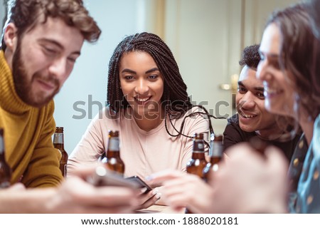 Friend`s gathering for feast. Cheerful youth is sharing positive emotion watching smartphone screens and drinking beers. Multiracial young people having fun drinking beers and using social media app
