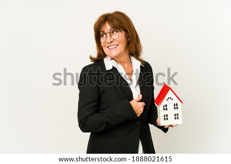Middle aged real estate agent holding a house model isolated looks aside smiling, cheerful and pleasant.