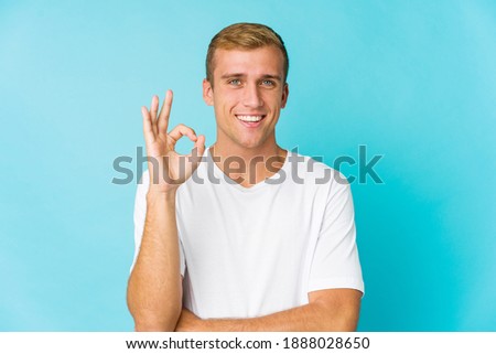 Young caucasian handsome man winks an eye and holds an okay gesture with hand.