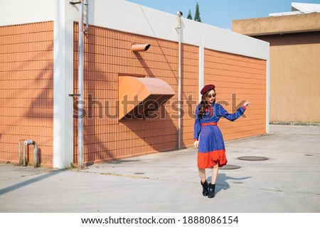 Wavy brunette woman in blue and red retro dress walking alone on roof top in sunlight.