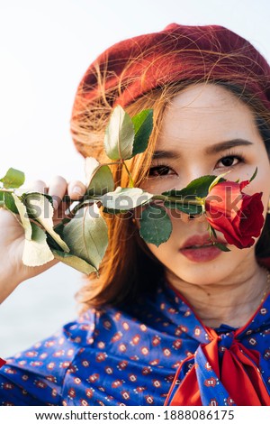 Close up shot of wavy brunette woman in blue and red retro dress hide her face with the rose.
