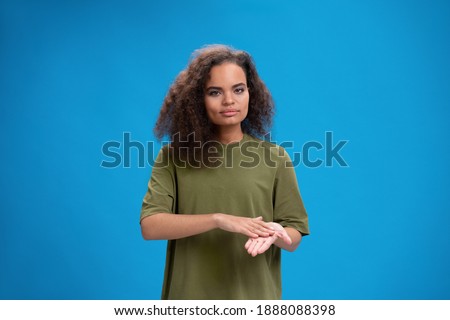 Deaf African American girl shows hand gesture using sign language. Young woman positively looking at camera wearing in an olive t-shirt. Isolated on blue background. Beauty concept. Skin care, hand