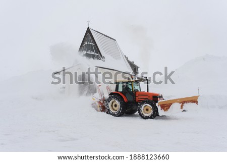 Snow blower shooting snow into air to clear road after heavy snowfall in winter landscape with tiny church steeple in background