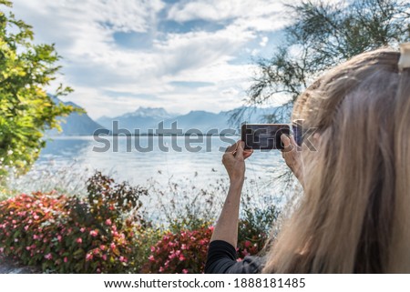 Photo with copy space of the hands of a woman taking a photo with the mobile of an alpine lake surrounded by snowy peaks. Montreux, Lake Geneva in Switzerland