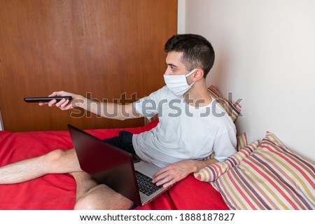 Bored man in bed wearing a face mask and watching television 