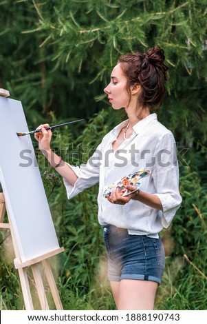 Girl artist, woman paints a picture, summer forest, white shirt denim shorts, creating creativity and artistic mood. First brush stroke, getting started. Background of green grass trees and spruce