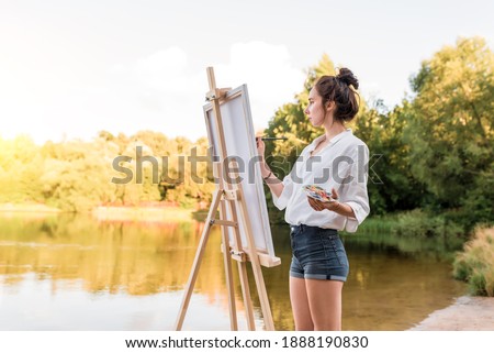 girl artist, woman paints picture of landscape, summer lake river pond, white shirt denim shorts, creating creativity artistic. Blank white canvas. Background trees water. Brush color palette paints