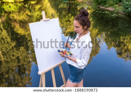 woman draws picture, girl artist, summer in park lake pond river, shirt shorts, creating creativity artistic mood. Blank white canvas, getting started. Water background. Brush color palette paints.