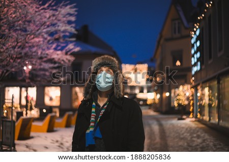 the attractive kind man in a fur hat and face mask with illuminated city lights  at night during the celebration of Christmas and on New Year's Eve 2021 during the coronavirus covid-19 pandemic
