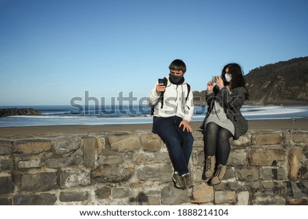 A young man looking at his mobile phone while a young woman takes a picture of him on her phone