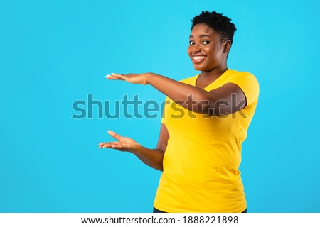 Cheerful Overweight African American Woman Holding Invisible Object Advertising Your Product Or Showing Size Standing Posing Over Blue Studio Background, Smiling To Camera. Copy Space