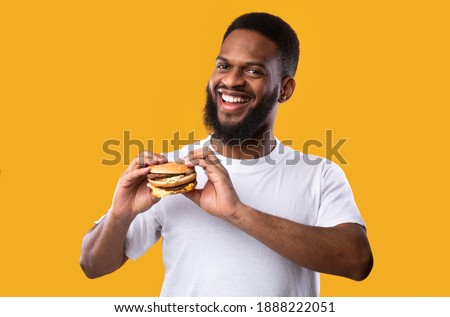 Junk Food. Happy African American Guy Posing With Burger Standing On Yellow Studio Background, Smiling To Camera. Nutrition And Overeating Habit, Fast Food Lover Concept