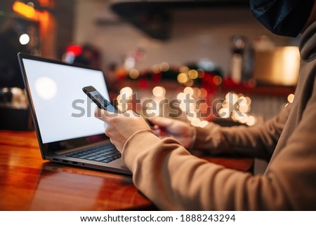 Shopping and online payment using cellphone, laptop and credit card. Man wearing mask during pandemic quarantine sits in a cafe and pays for goods via internet. New normal and technologies concept