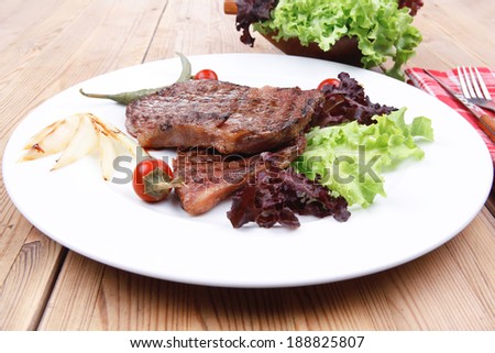 meat food : two grilled steak on green lettuce salad , with roast onion and red hot chili peppers , on dish over wooden table