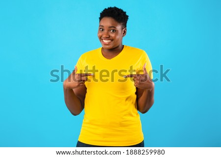 Choose Me. Cheerful Oversized African Lady Pointing Fingers At Herself Smiling To Camera Standing Posing Over Blue Background In Studio. I'm The Best, Self-Confidence Concept