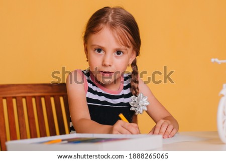 Little girl draws on a white table with crayons. Creative child. Studio photo on a yellow background.