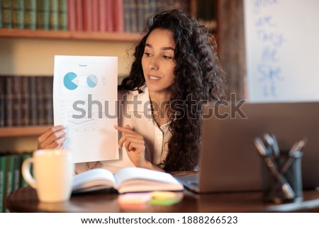 Portrait of female financial analyst having video call on laptop, giving virtual presentation. Woman showing statistics, graphs and charts to camera, holding document paper, explaining report