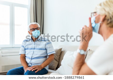 Senior friends having conversation on the sofa at home during Covid 19 pandemic. Elderly wearing medical face mask, preventing covid 19 coronavirus pandemic infection spread. Social distancing