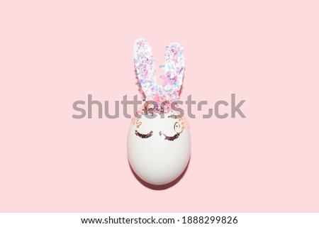 cute Easter bunny shape egg with eyes and shiny glitter and confetti ears on a pastel pink background with selective focus